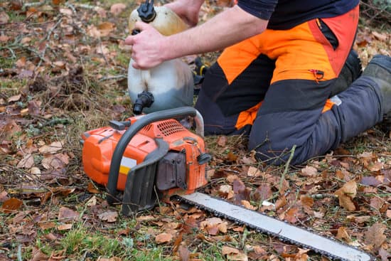 A close-up of an orange chainsaw on fallen leaves with a worker in orange safety trousers refuelling it.