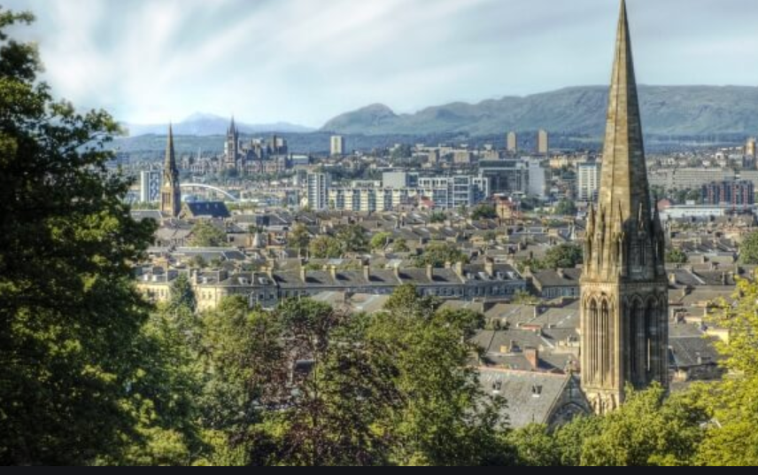 A view over a city skyline with a tall stone church spire, rooftops, and hills in the distance, framed by tree branches in the foreground.