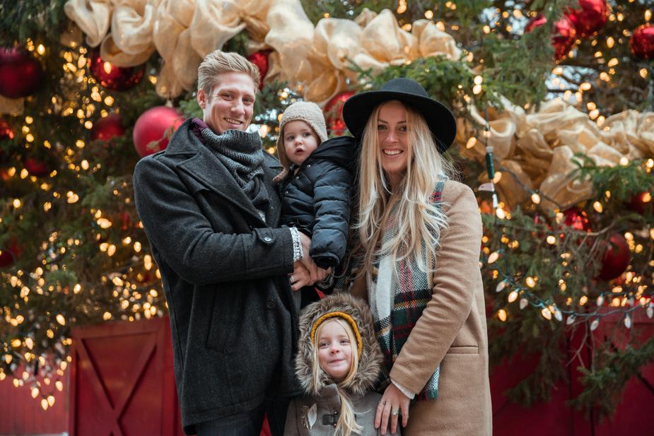 A family of four in winter coats smiles together in front of a Christmas tree decorated with lights, red baubles and gold ribbon.