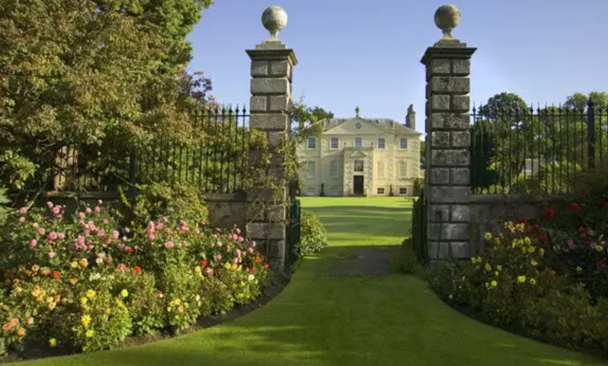 A large stone country house seen through ornate gate pillars, with lawn and colourful flower borders in the foreground.