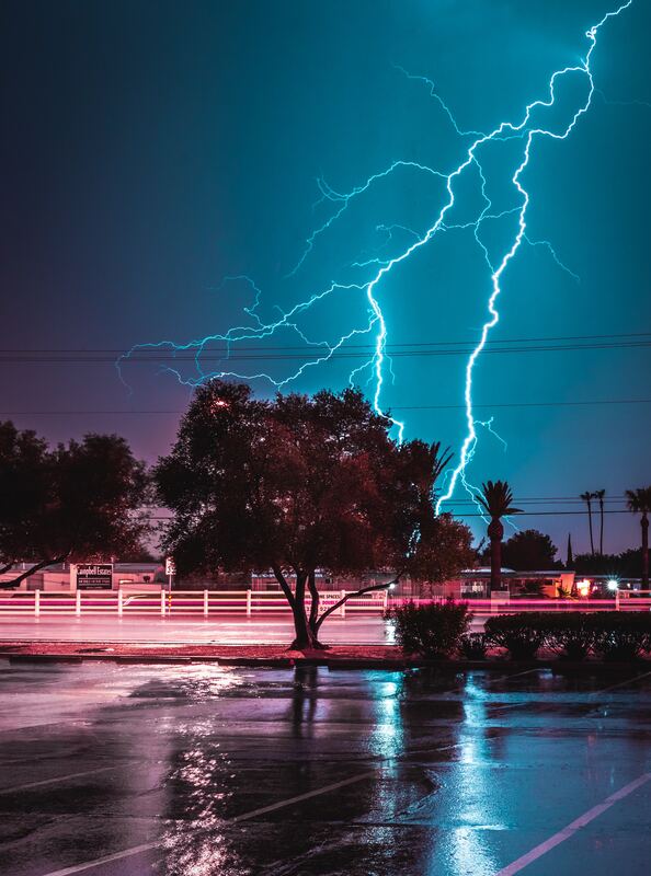 Bright lightning bolts strike the sky above a tree and a rain-soaked car park at night.