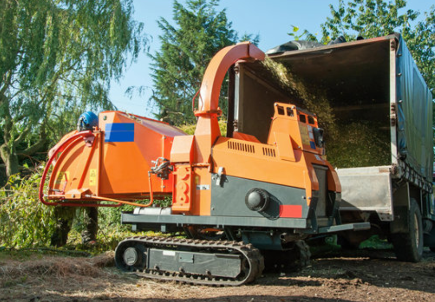 An orange tracked wood chipper machine blowing wood chips into the back of a truck near felled branches.