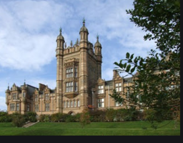 A large ornate sandstone Victorian building with turrets and towers, seen behind a tree and lawn.