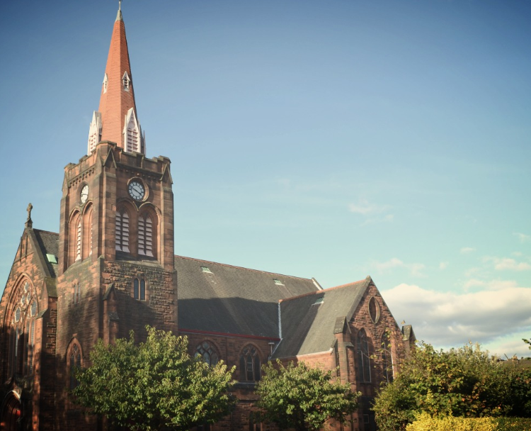 A red sandstone church with a tall spire and clock tower rising above surrounding trees.