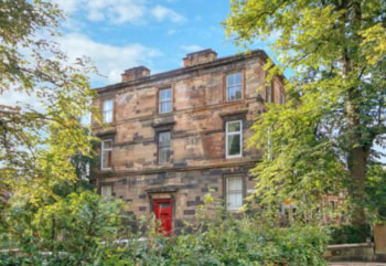 A three-storey sandstone tenement building with a red front door, surrounded by trees and greenery.