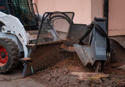 A skid steer loader fitted with a stump grinder attachment cutting into a tree stump beside a building wall.