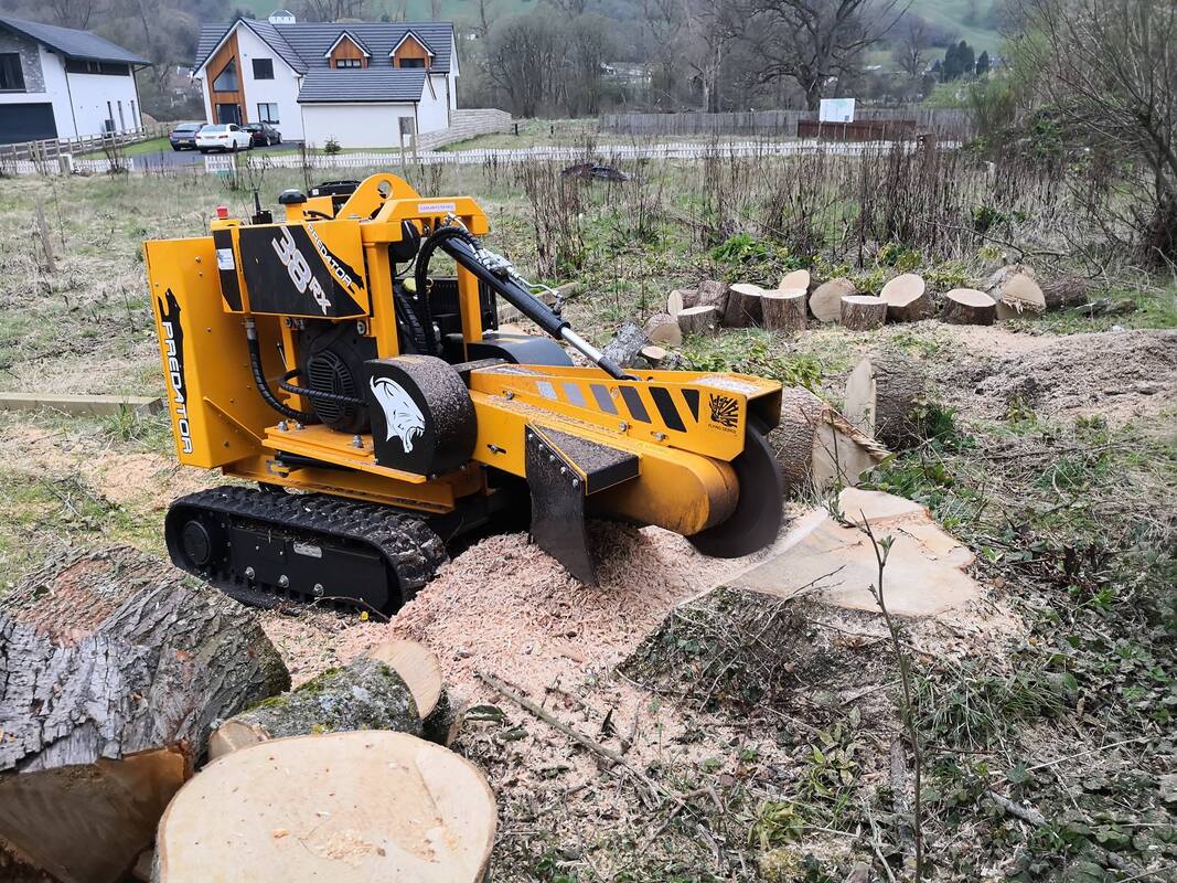 A yellow tracked stump grinder machine grinding a large tree stump in front of houses with cut log rounds stacked nearby.